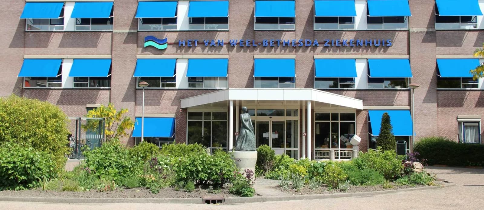 Hoofdingang van Het Van Weel-Bethesda Ziekenhuis in Dirksland met blauwe zonneschermen en een beeld voor de deur