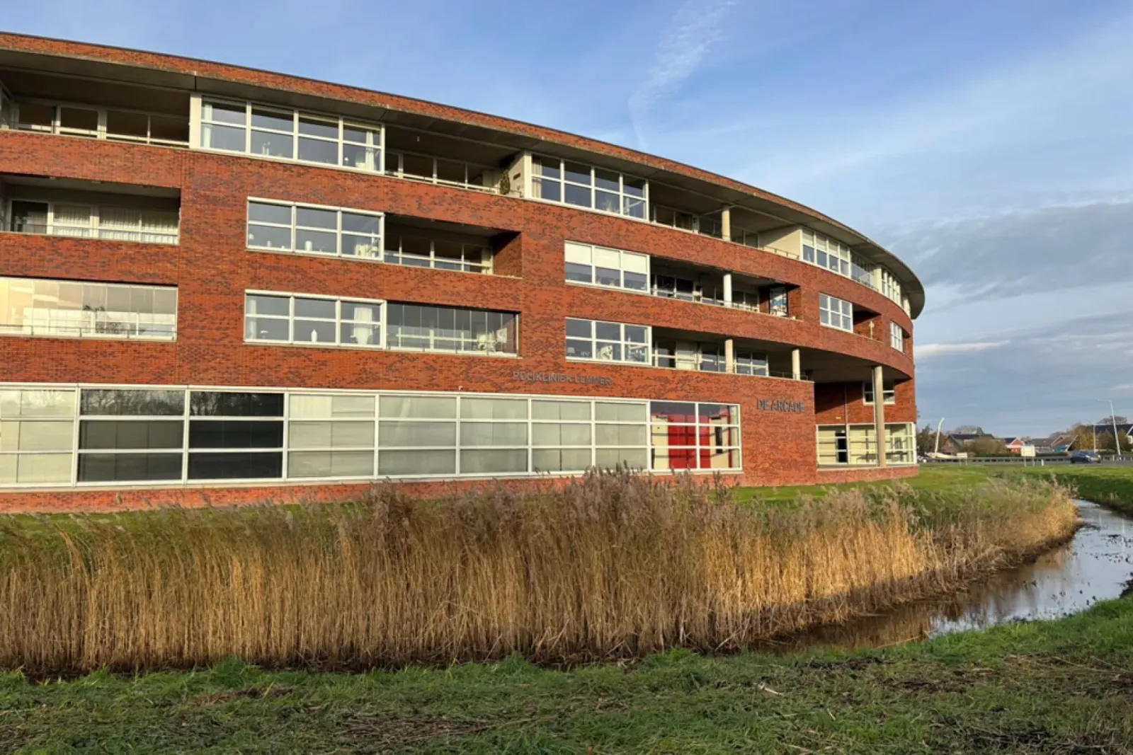 Rond bakstenen woon- en bedrijvencomplex De Arcade aan het water, met de tekst Polikliniek Lemmer op de gevel