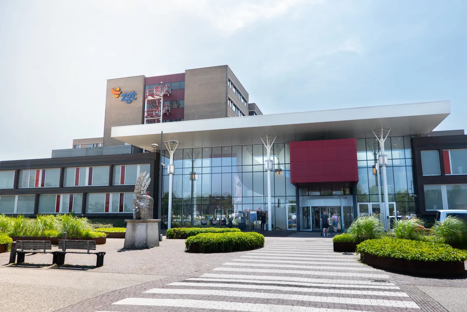 Hoofdingang van ZGT Almelo met glazen gevel en rode luifel, gezien vanaf het voorplein