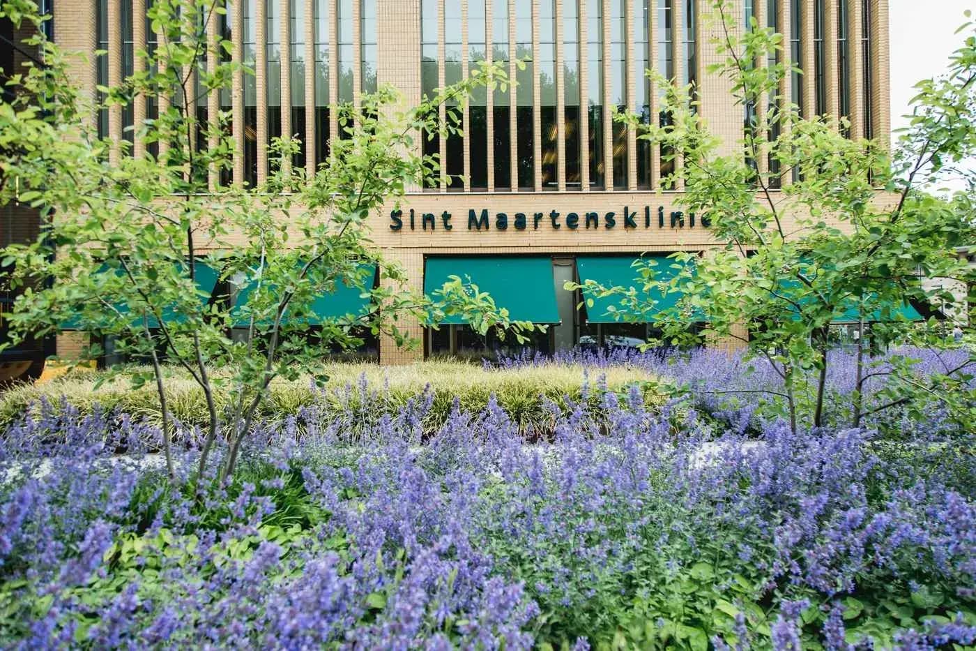 Gevel van de Sint Maartenskliniek in Nijmegen met de naam boven de ingang en bloeiende planten ervoor